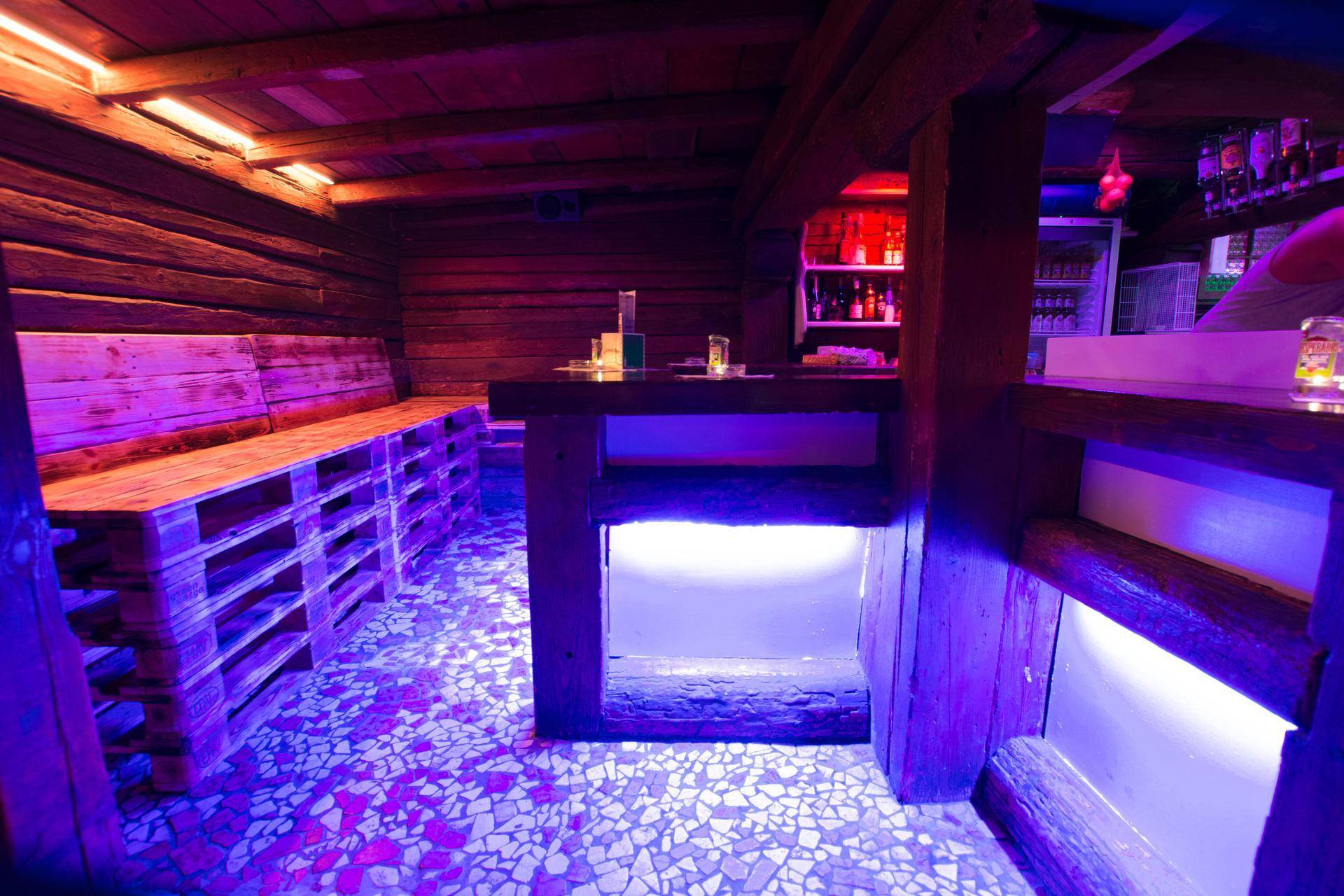 Barbereich mit beleuchteter Theke, Holzbänken und Palettenmöbeln in violetter Lichtstimmung.