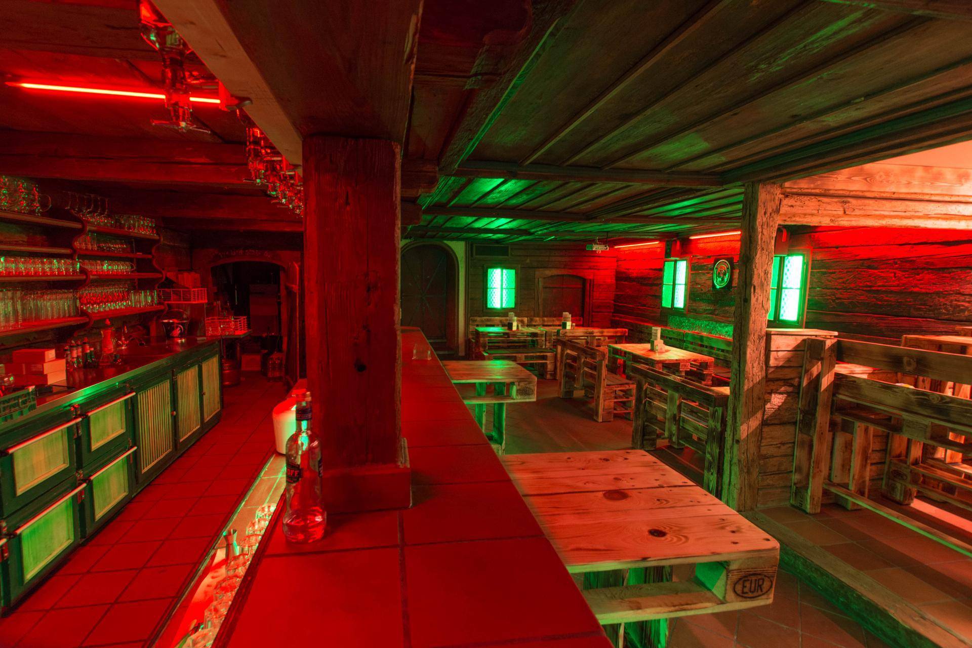 Weitläufiger Barbereich mit Holzbalken, Palettenmöbeln und einer Mischung aus roter und grüner Beleuchtung.