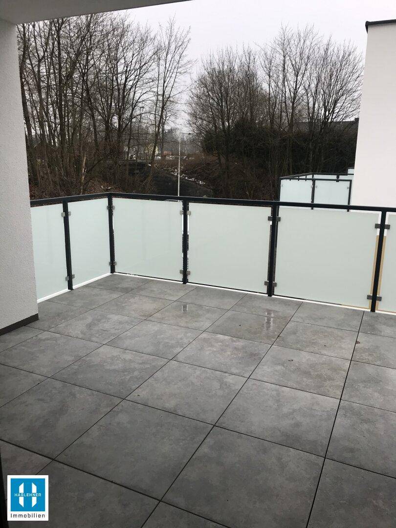 Geräumiger Balkon mit grauen Fliesen und Milchglasgeländer, umgeben von Bäumen.