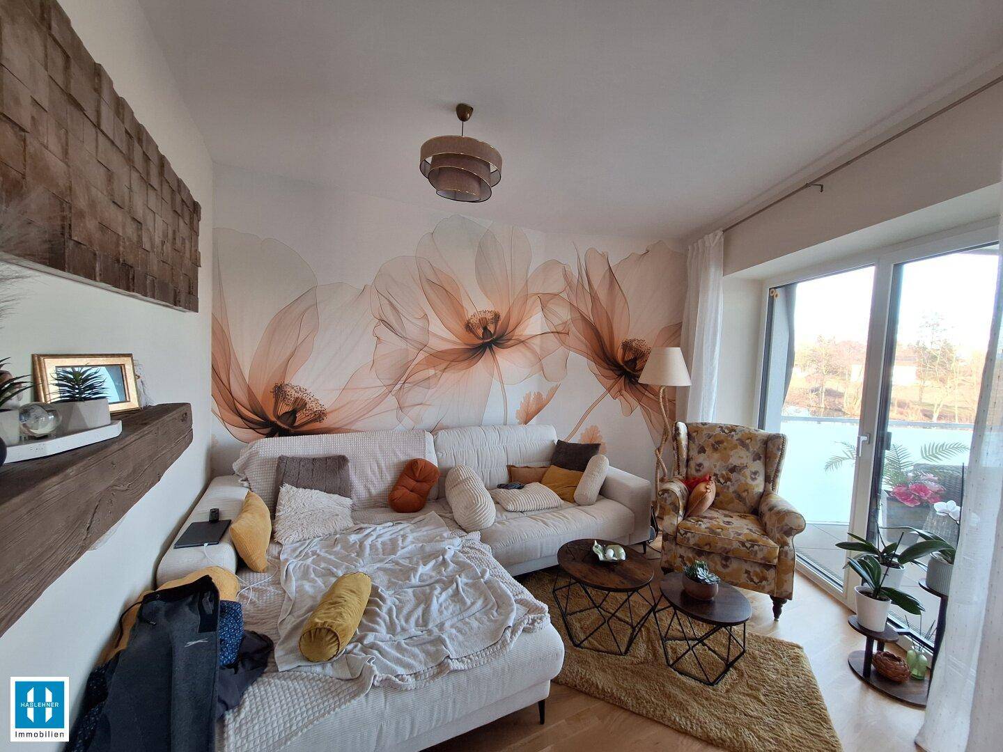 Helles Wohnzimmer mit bequemer Couch, floraler Wandtapete und Zugang zum Balkon.