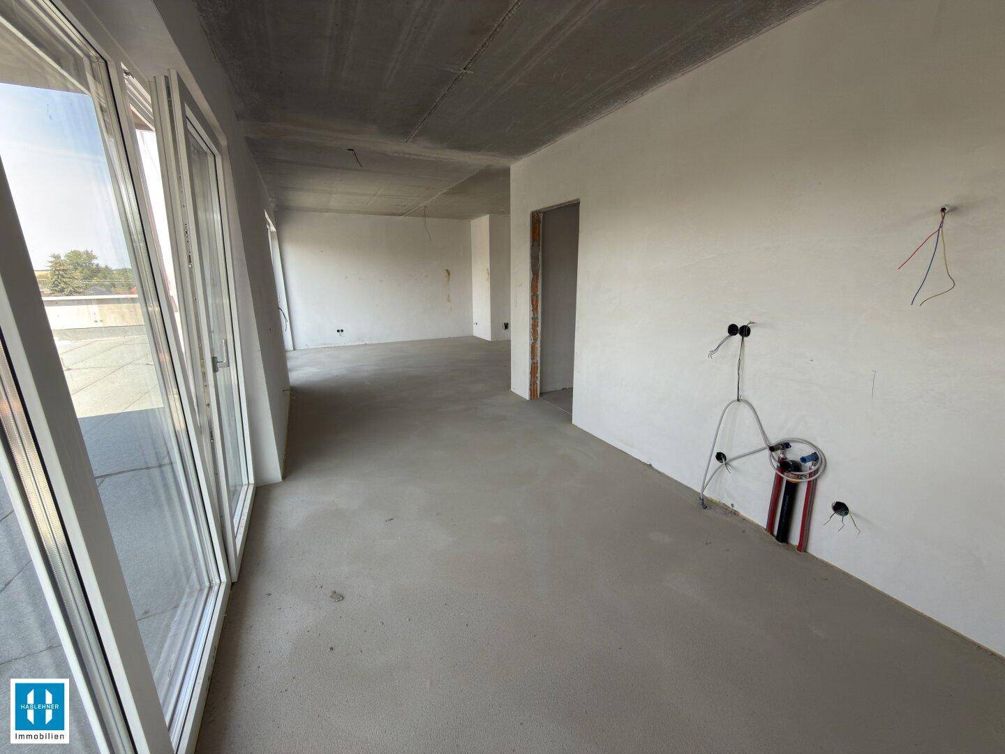 Unfertiger Innenraum mit Betonboden, großen Fenstern und sichtbaren Sanitärinstallationen an der Wand.