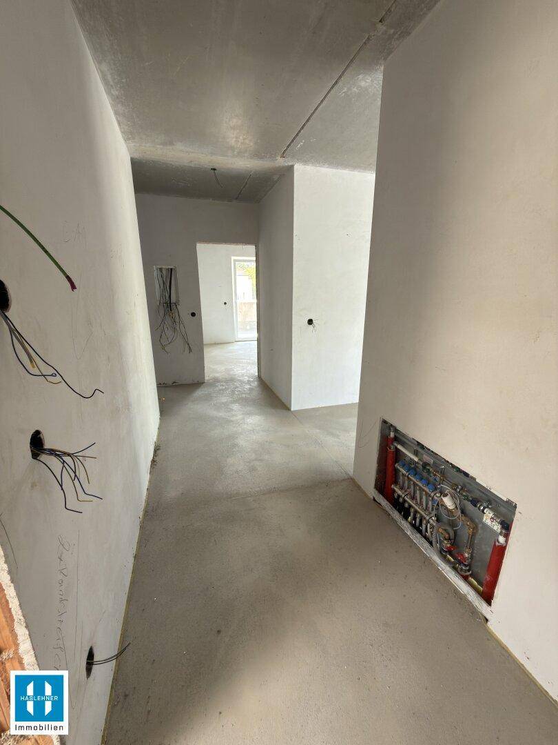 Unfertiger Flur mit Betonboden, sichtbaren Leitungen und einer Heizungsverteilung in der Wand.