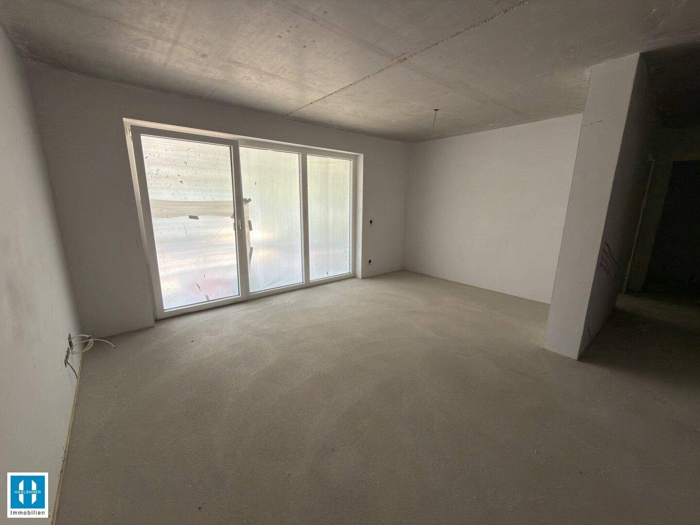 Heller Rohbau-Raum mit großen Fenstern, die viel Tageslicht hereinlassen, und Betonboden.