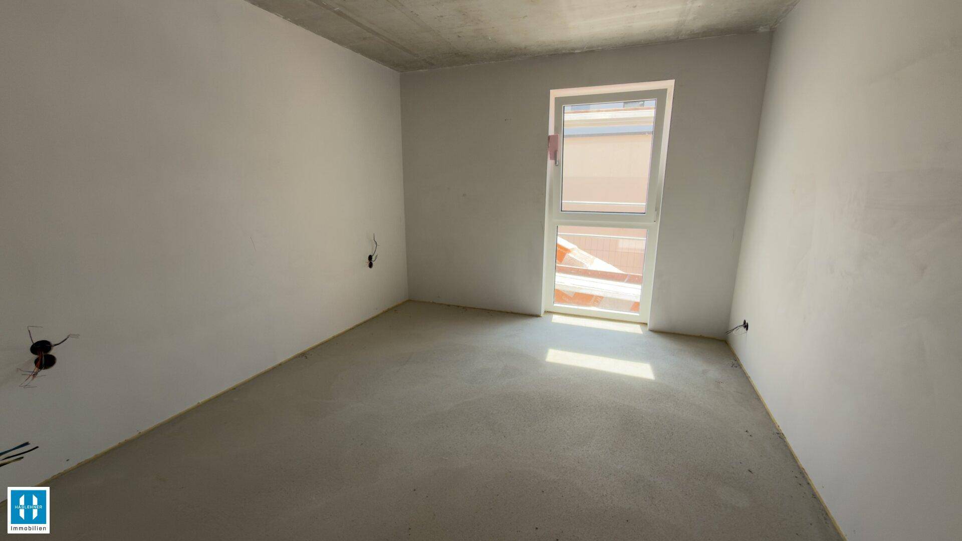 Unfertiger Raum mit Betonboden und einem Fenster, das viel natürliches Licht hereinlässt.