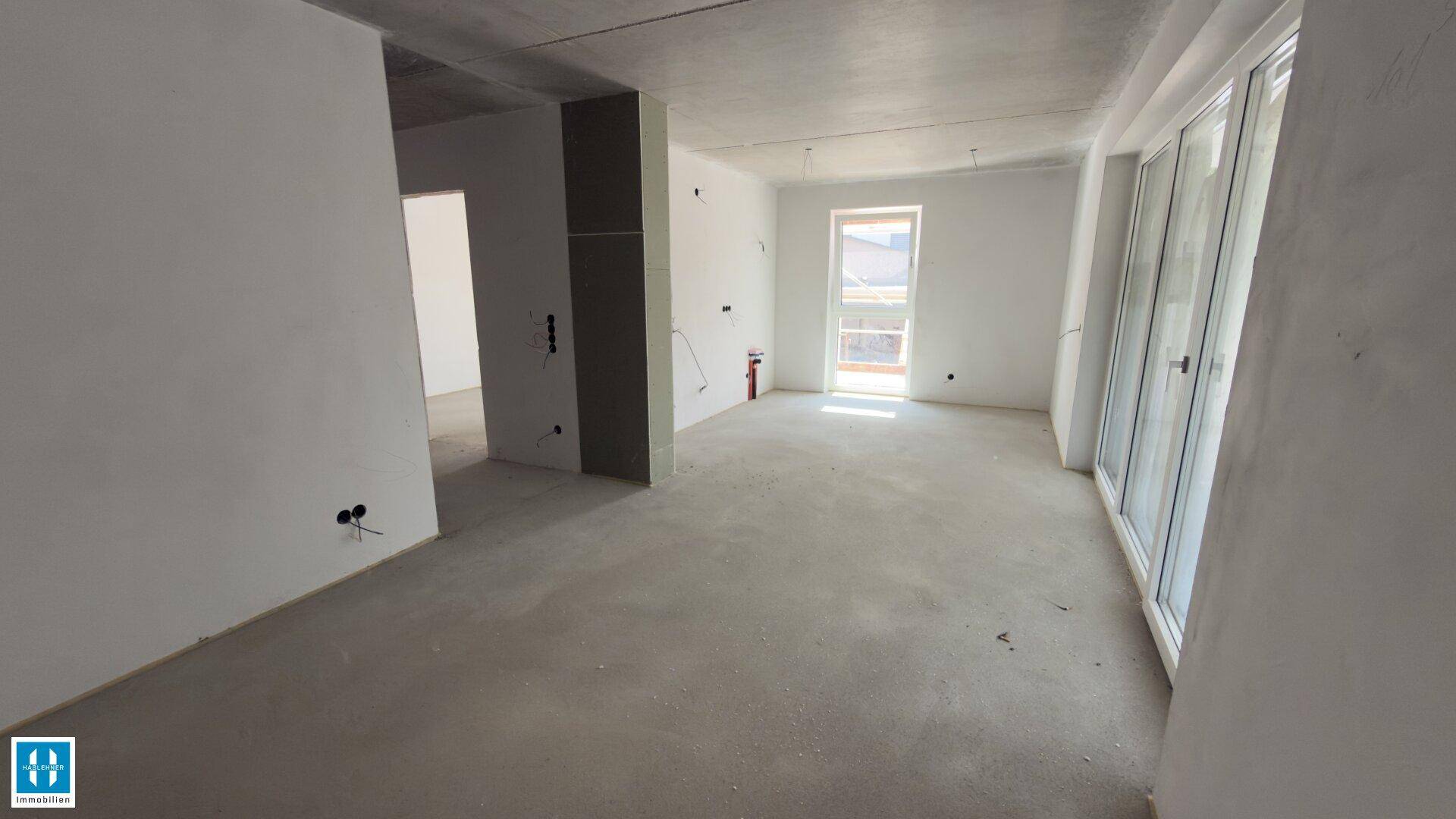 Geräumiger, unfertiger Innenraum mit Betonboden und großen Fenstern, bereit für den Ausbau.