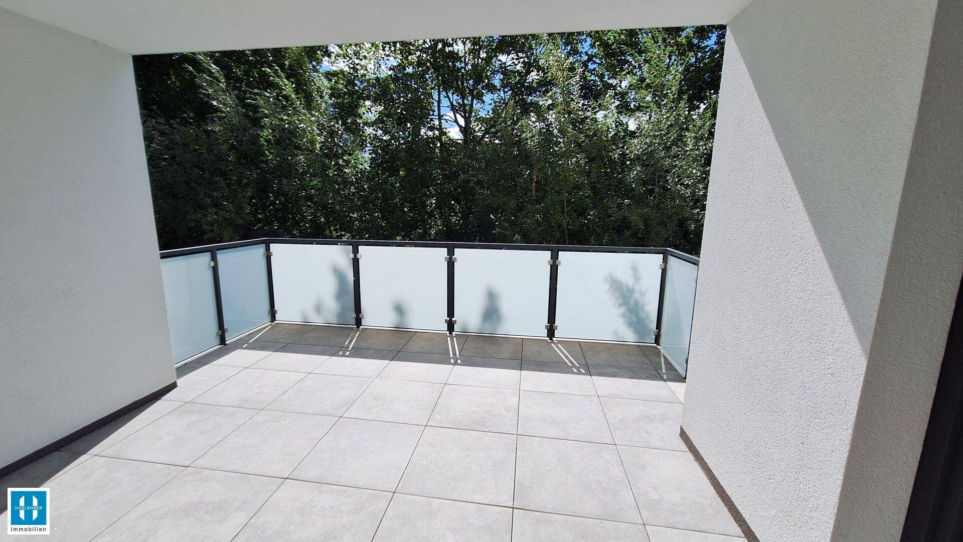 Geräumiger Balkon mit Fliesenboden und Glasgeländer, umgeben von Bäumen und Natur.