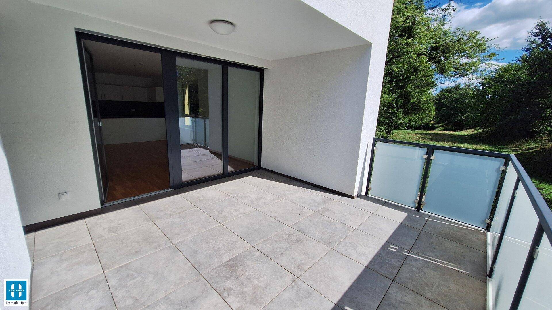 Heller Balkon mit Fliesenboden und Zugang zum Innenraum, umgeben von viel Grün.