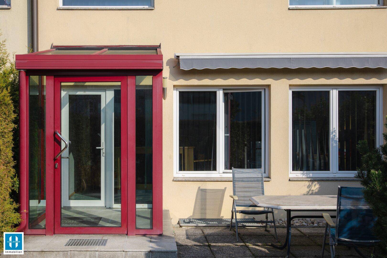 Überdachter Eingangsbereich mit rotem Windfang und angrenzender Terrasse mit Gartenmöbeln.