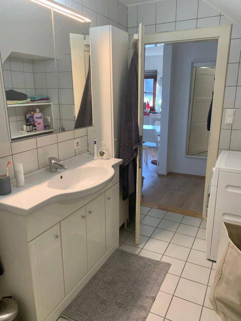 Badezimmer mit Waschbecken, Spiegelschrank und Waschmaschine, mit Blick in den Essbereich.