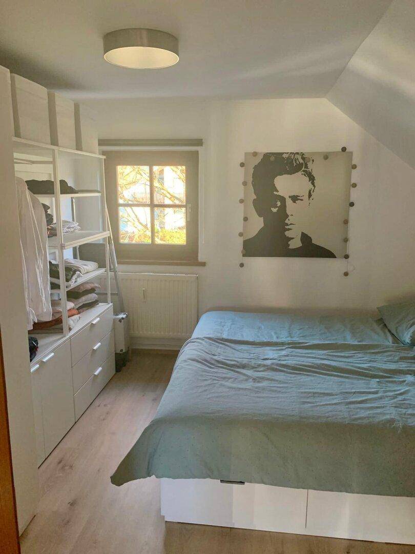 Schlafzimmer mit Bett, Kleiderschrank und Fenster, das Tageslicht hereinlässt.