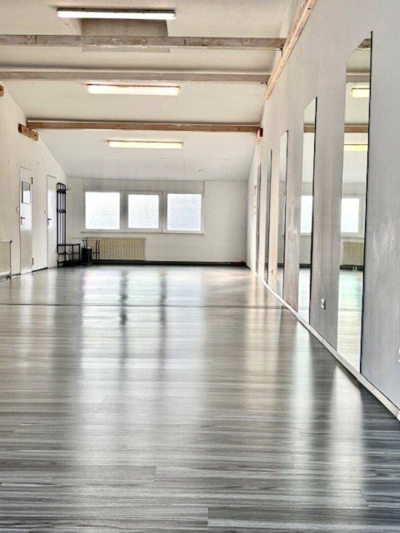 Langer, offener Raum mit glänzendem Laminatboden, großen Fenstern und sichtbaren Deckenbalken.