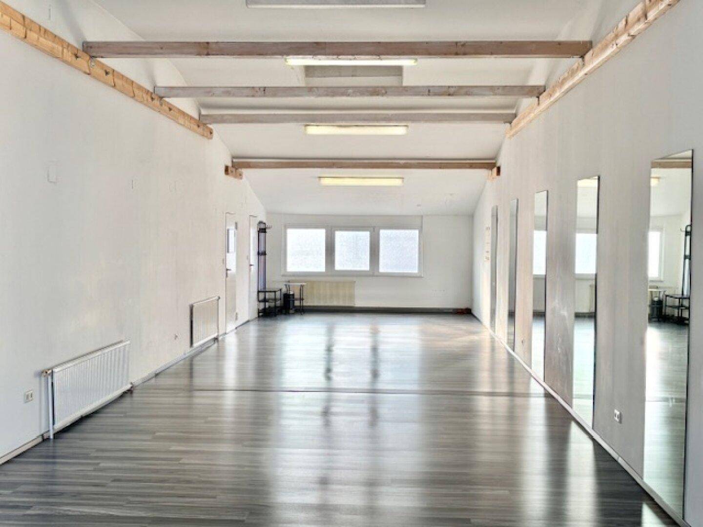 Weitläufiger, heller Gewerberaum mit Holzdeckenbalken, Laminatboden und großen Fenstern.