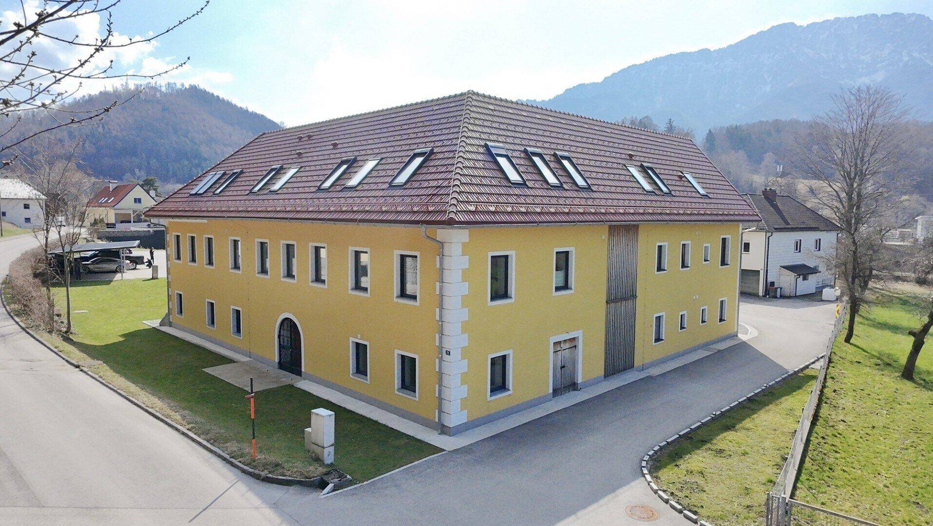 Großes gelbes Gebäude mit rotem Ziegeldach und vielen Dachfenstern, umgeben von Natur und Bergen.