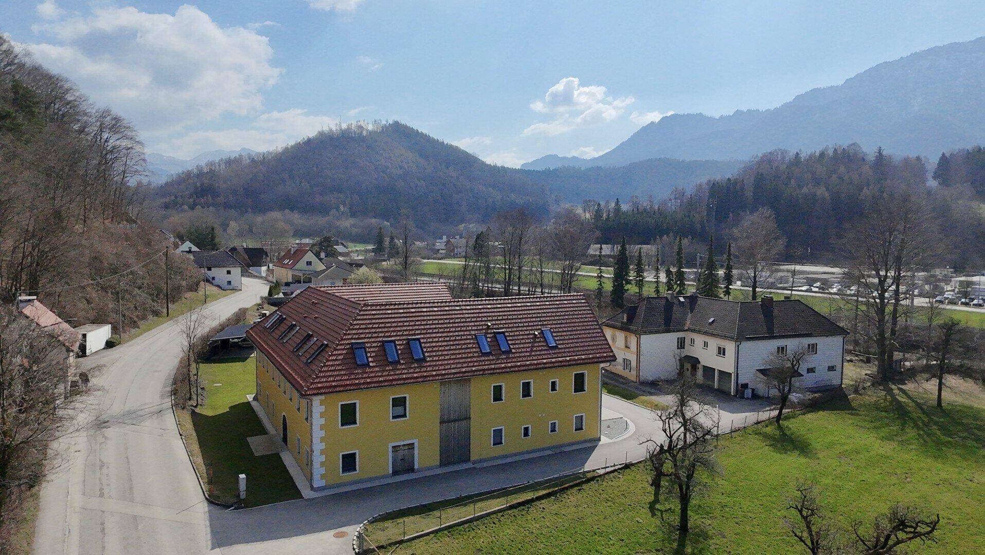 Luftaufnahme eines gelben Gebäudes mit rotem Dach, eingebettet in eine grüne Landschaft mit Bergen.