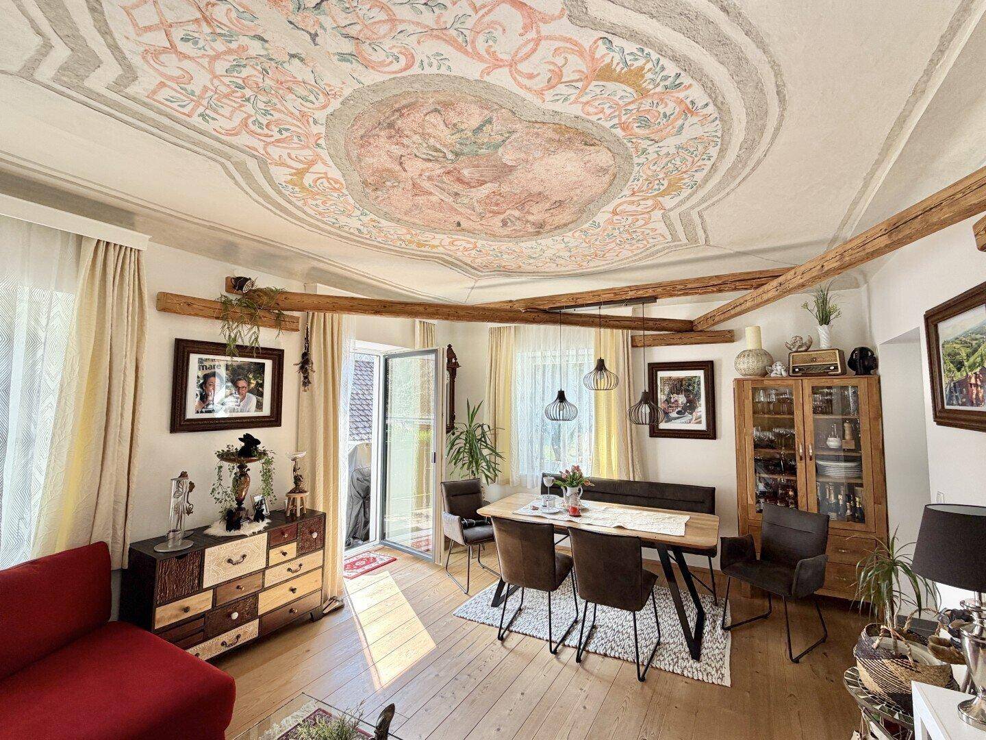 Gemütlicher Wohn-Essbereich mit kunstvoller Deckenmalerei, Holzbalken und Zugang zum Außenbereich.