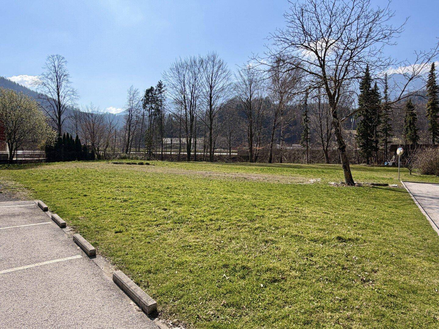 Große, grüne Rasenfläche neben dem Gebäude mit Parkplätzen und Bäumen im Hintergrund.