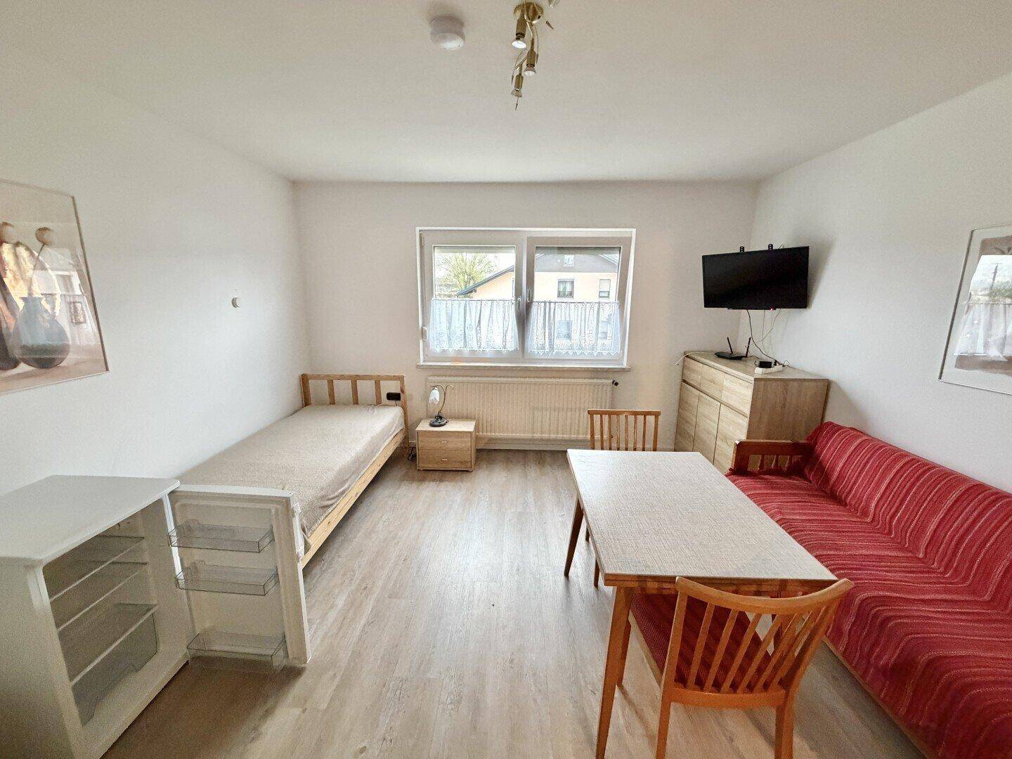 Einfaches Zimmer mit Einzelbett, Esstisch und kleinem Kühlschrank.
