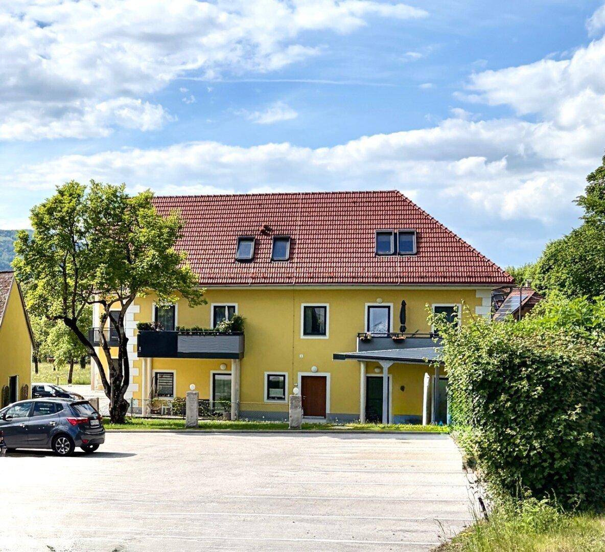 Ansicht des gelben Wohnhauses mit rotem Ziegeldach, Balkonen und gepflegter Außenanlage unter blauem Himmel.