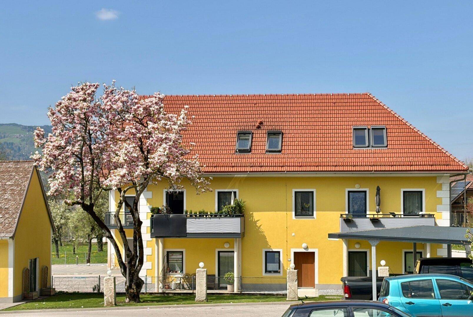 Gelbes Mehrfamilienhaus mit rotem Ziegeldach, Balkonen und blühendem Baum im Vordergrund an einem sonnigen Tag.