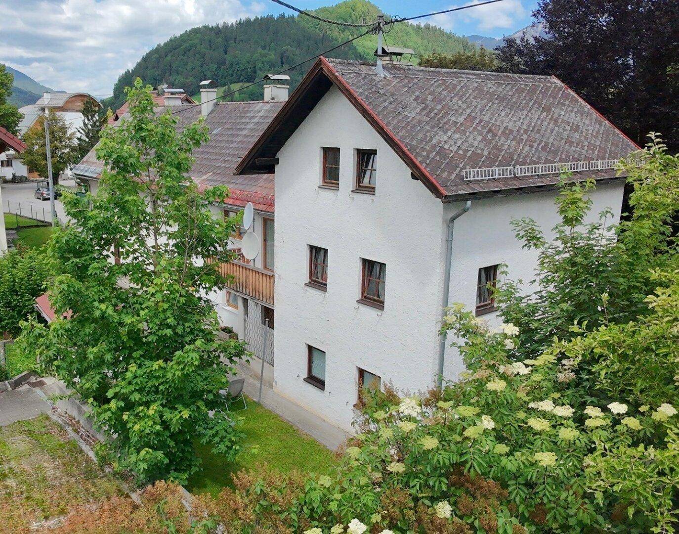 Seitenansicht des Gebäudes mit Satteldach, umgeben von üppigem Grün und Bäumen.