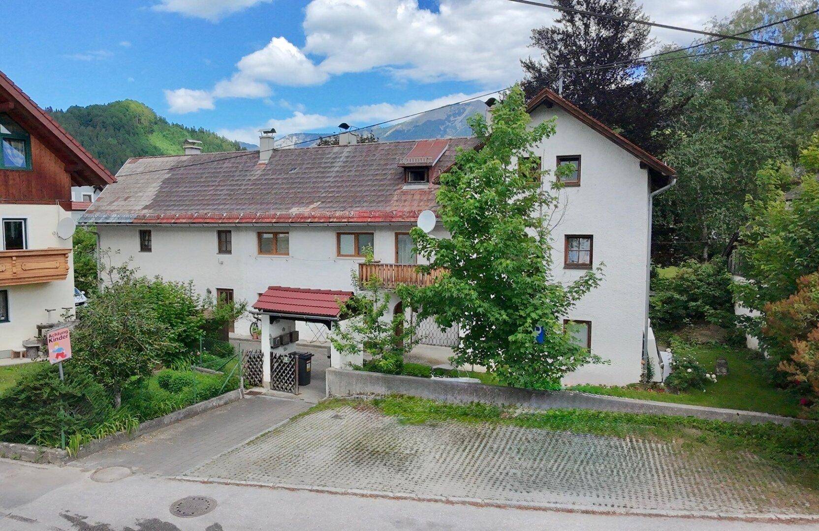 Anwesen mit Parkplatz, Garten und einem weiten Blick auf die umliegende Bergwelt unter blauem Himmel.