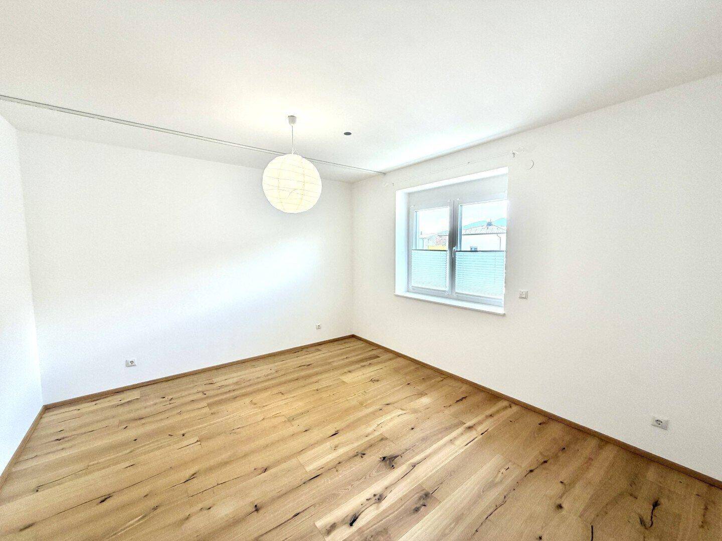 Leeres Schlafzimmer mit hellem Holzboden, weißen Wänden und einem Fenster mit Plissee.