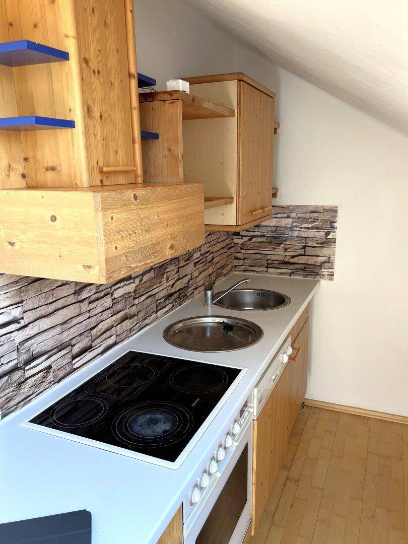 Kleine Küchenzeile mit Holzschränken, Steinoptik-Rückwand und Elektroherd unter einer Dachschräge.