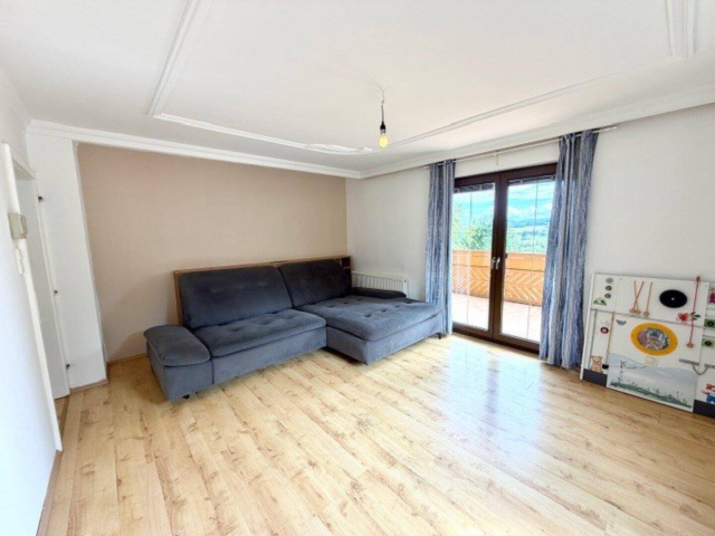 Gemütliches Wohnzimmer mit grauem Sofa, Laminatboden und direktem Zugang zum Balkon.