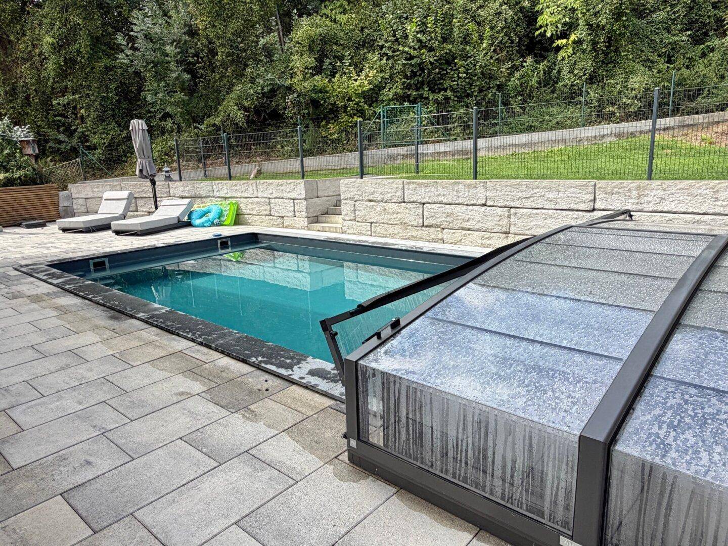 Außenpool mit verschiebbarer Abdeckung, umgeben von einer gepflasterten Terrasse und Liegestühlen.