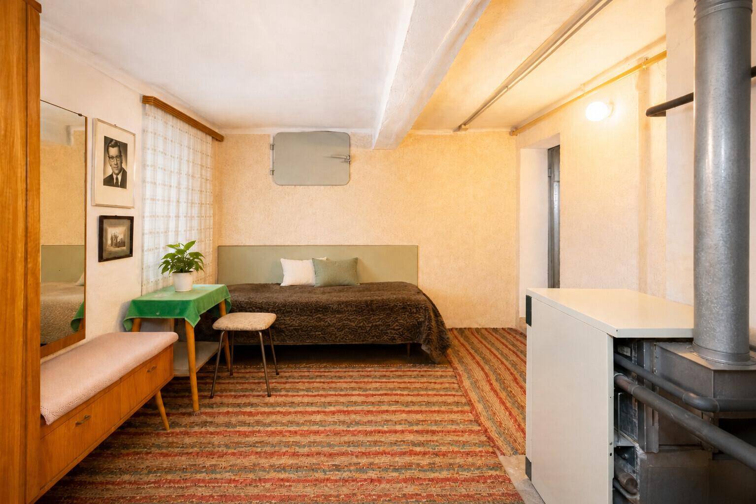Kellerraum mit Bett, Teppich und Heizungsrohren an der Decke.