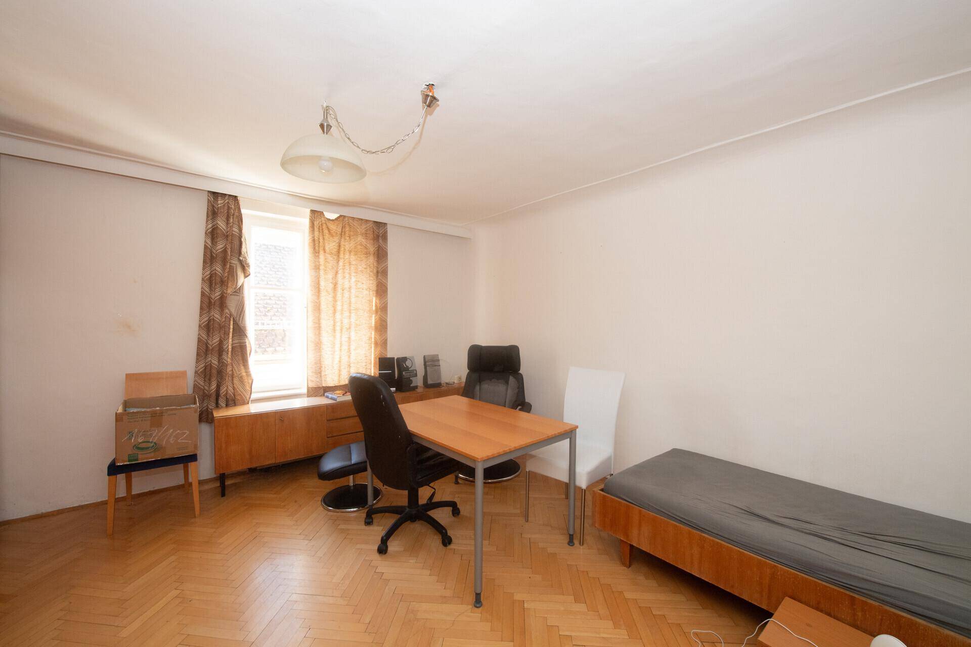 Einfaches Schlafzimmer mit Einzelbett, Schreibtisch, Parkettboden und einem Fenster mit Vorhängen.