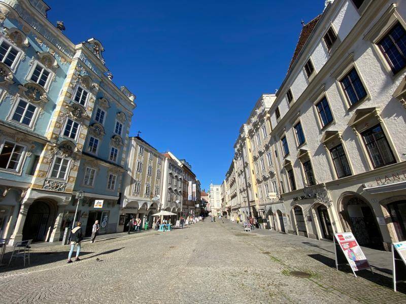 Weitläufige Straße mit historischen Gebäuden und Geschäften unter blauem Himmel, belebt mit Passanten.