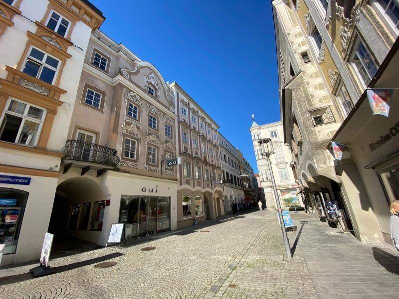 Historische Gebäudefronten mit Geschäften und einem Durchgang an einer gepflasterten Straße unter blauem Himmel.