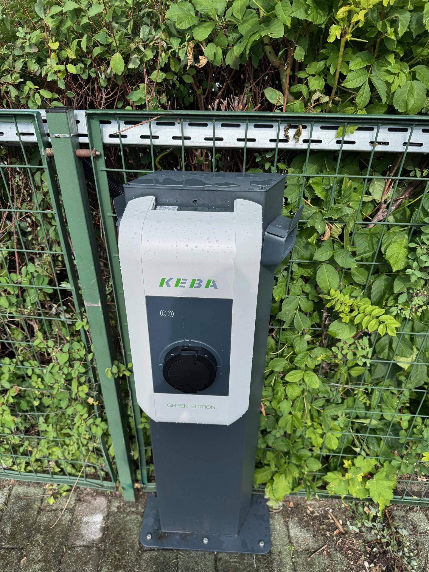 Eine KEBA Ladestation für Elektrofahrzeuge steht neben einem grünen Zaun mit dichtem Laubwerk.