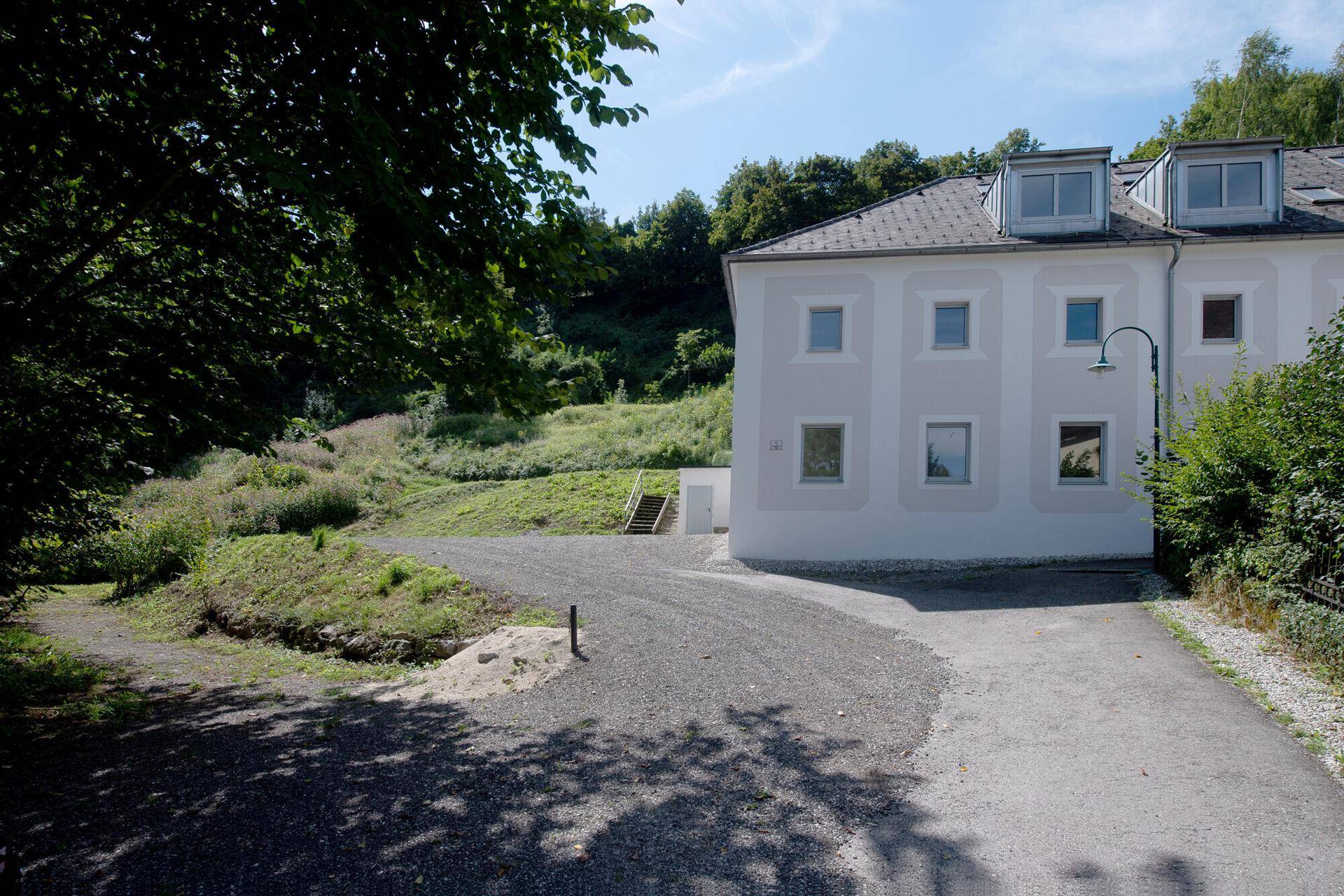 Seitenansicht des Hauses mit Zufahrt, Parkplatz und grüner, hügeliger Umgebung.