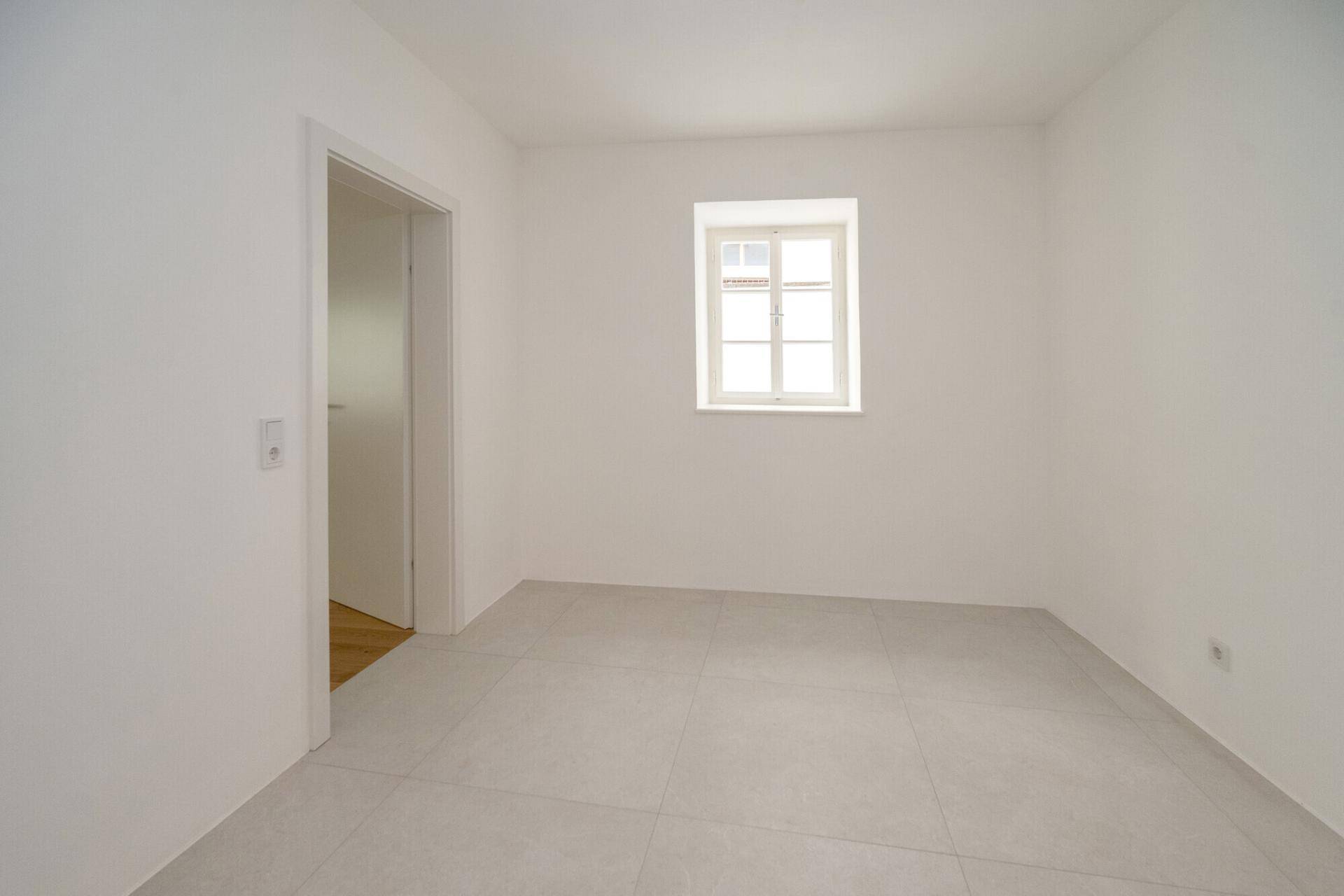 Leerer Raum mit hellgrauen Fliesen und einem Fenster, ideal für vielseitige Nutzung.