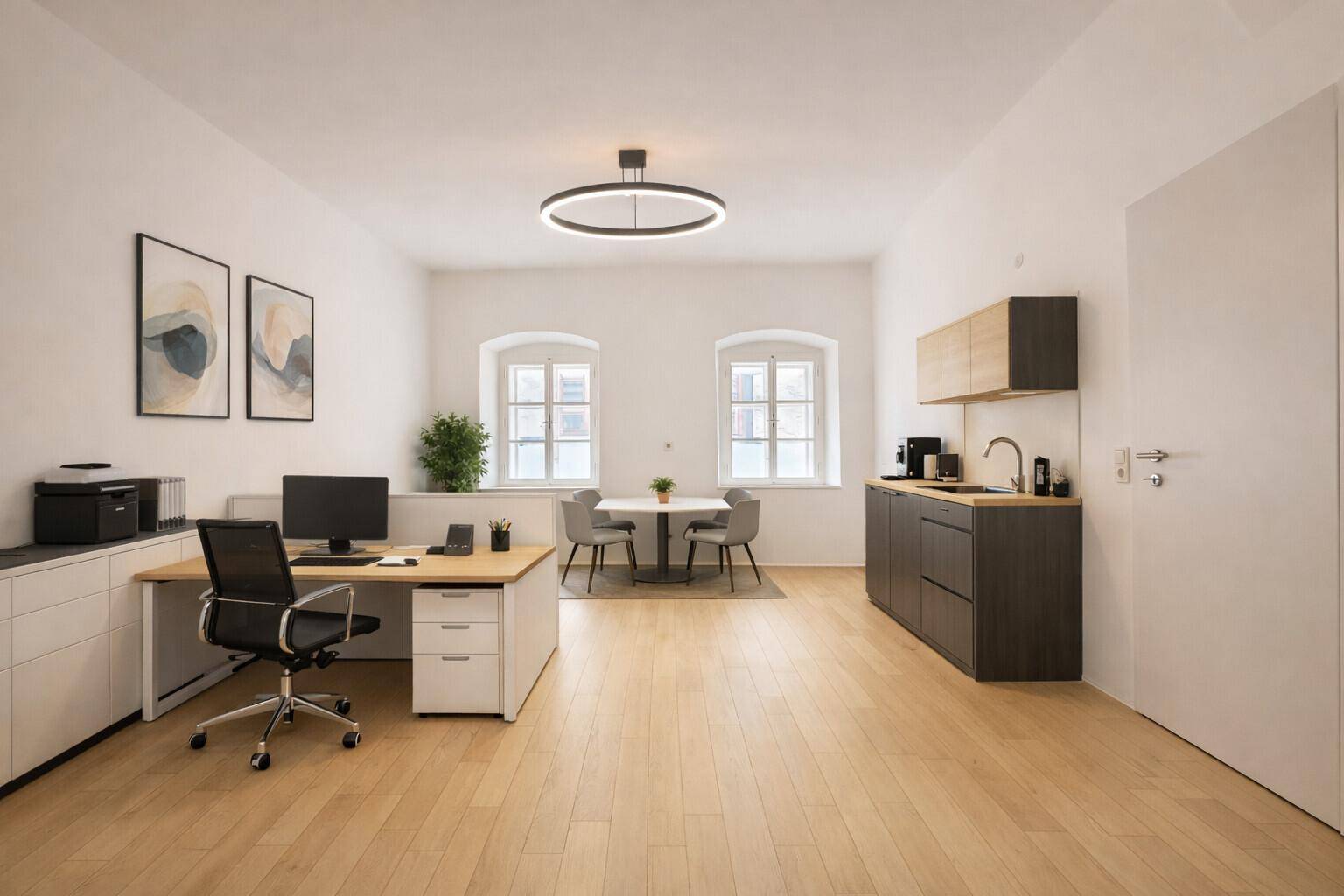Modern eingerichtetes Büro mit Holzboden, integrierter Küchenzeile und viel Tageslicht durch zwei Fenster.