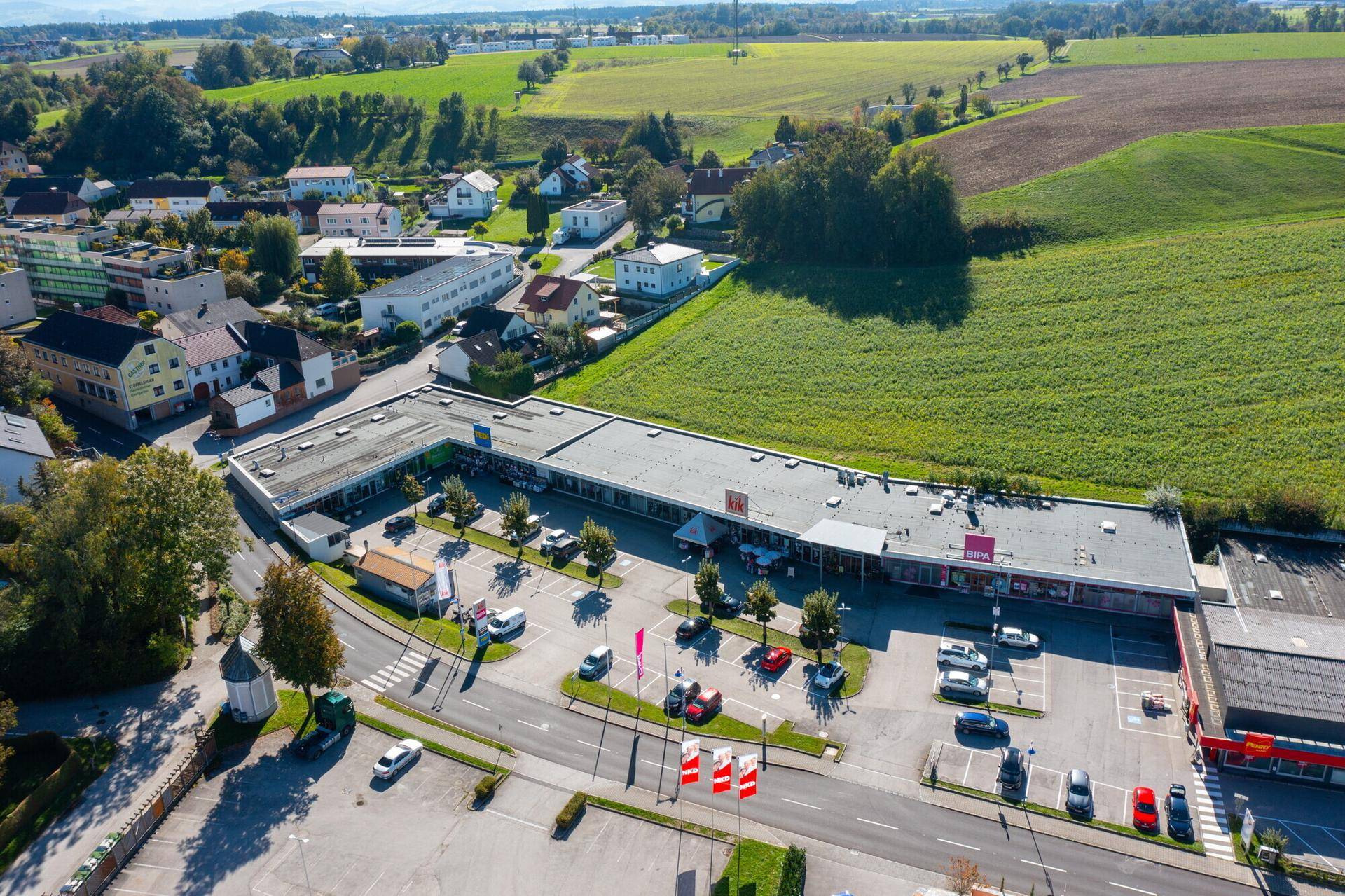 Luftaufnahme eines Gewerbegebiets mit Geschäften und Parkplätzen, angrenzend an Wohnbebauung und Natur.