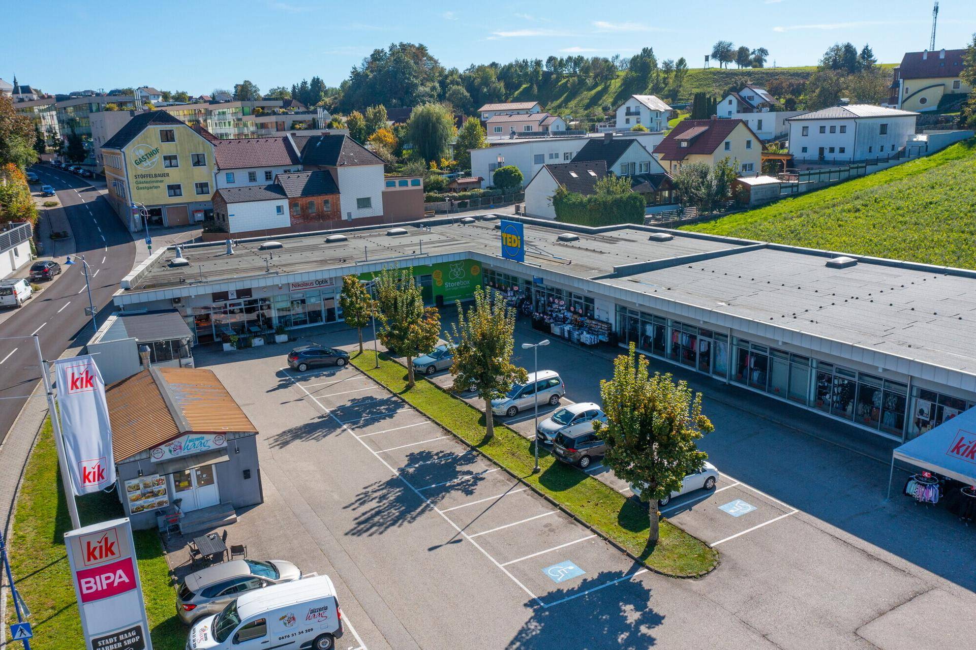Ansicht der Ladenfronten eines Einkaufszentrums mit davorliegendem Parkplatz und Bäumen.