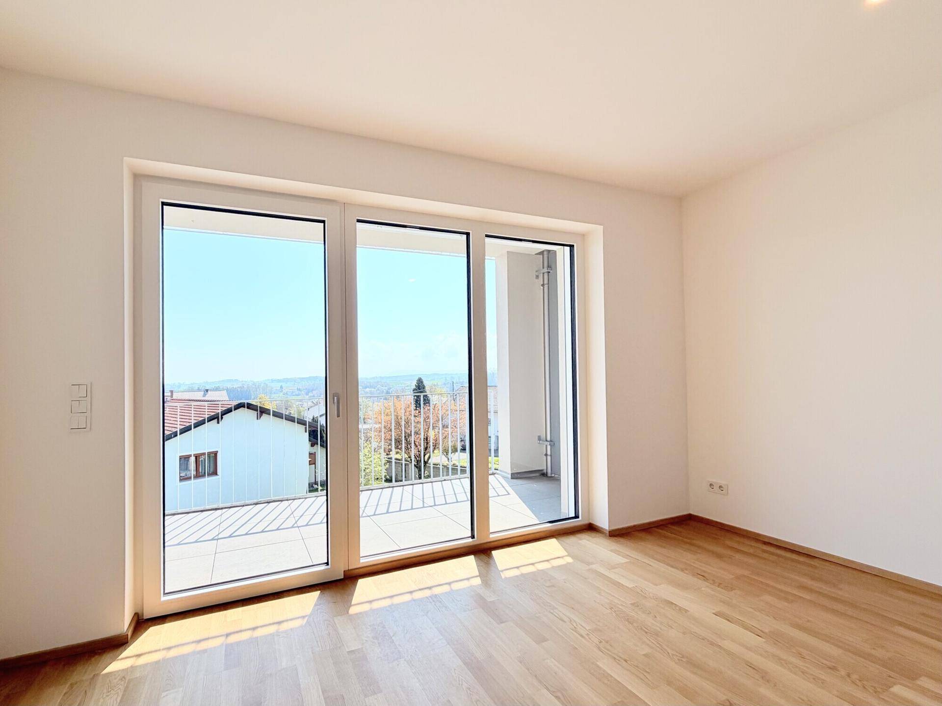 Helles Zimmer mit Holzboden und großem Fenster, das Zugang zu einem Außenbereich bietet.