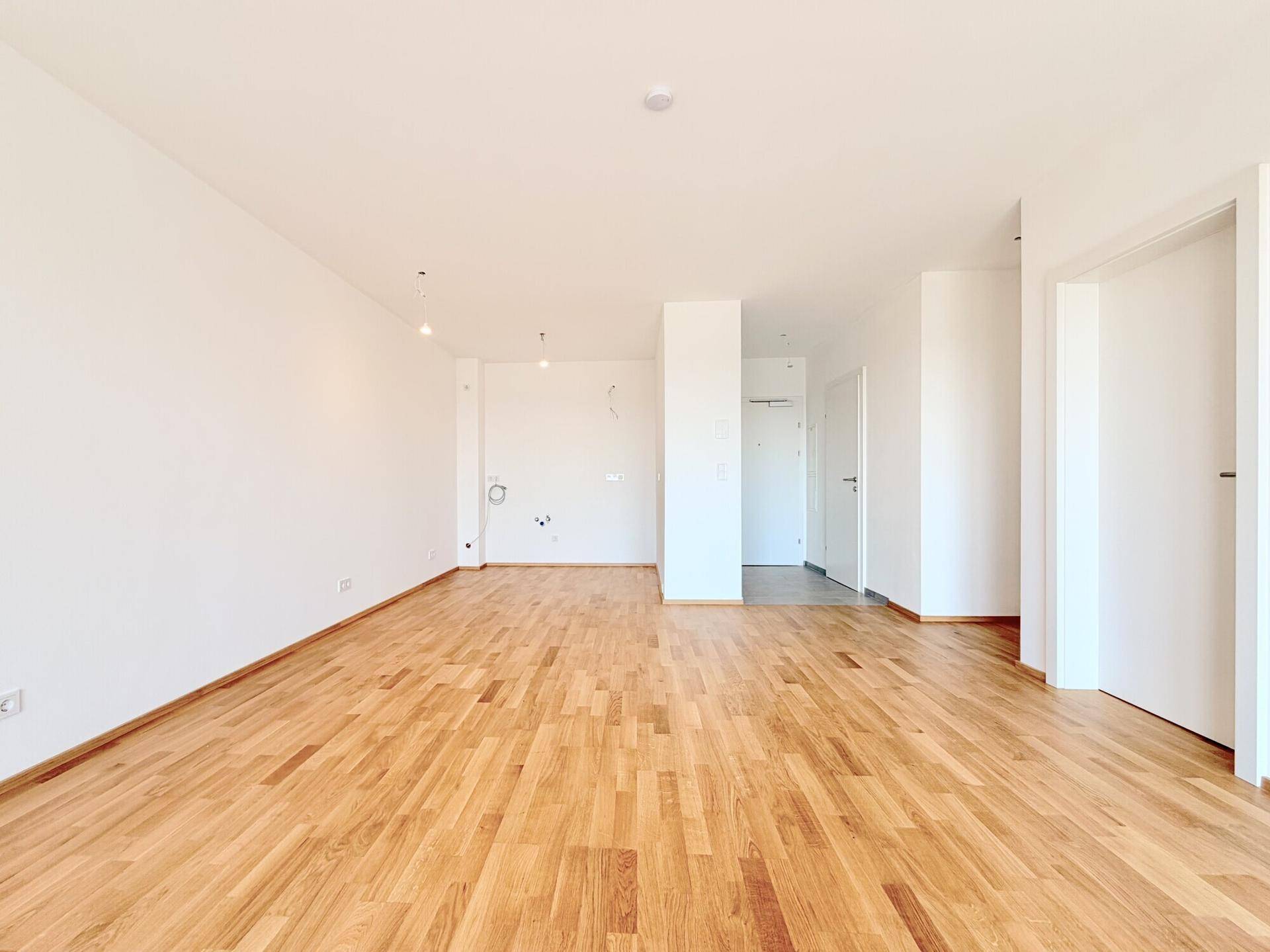 Heller Raum mit Holzboden und vorbereiteten Anschlüssen für eine Küche oder Mediengeräte.