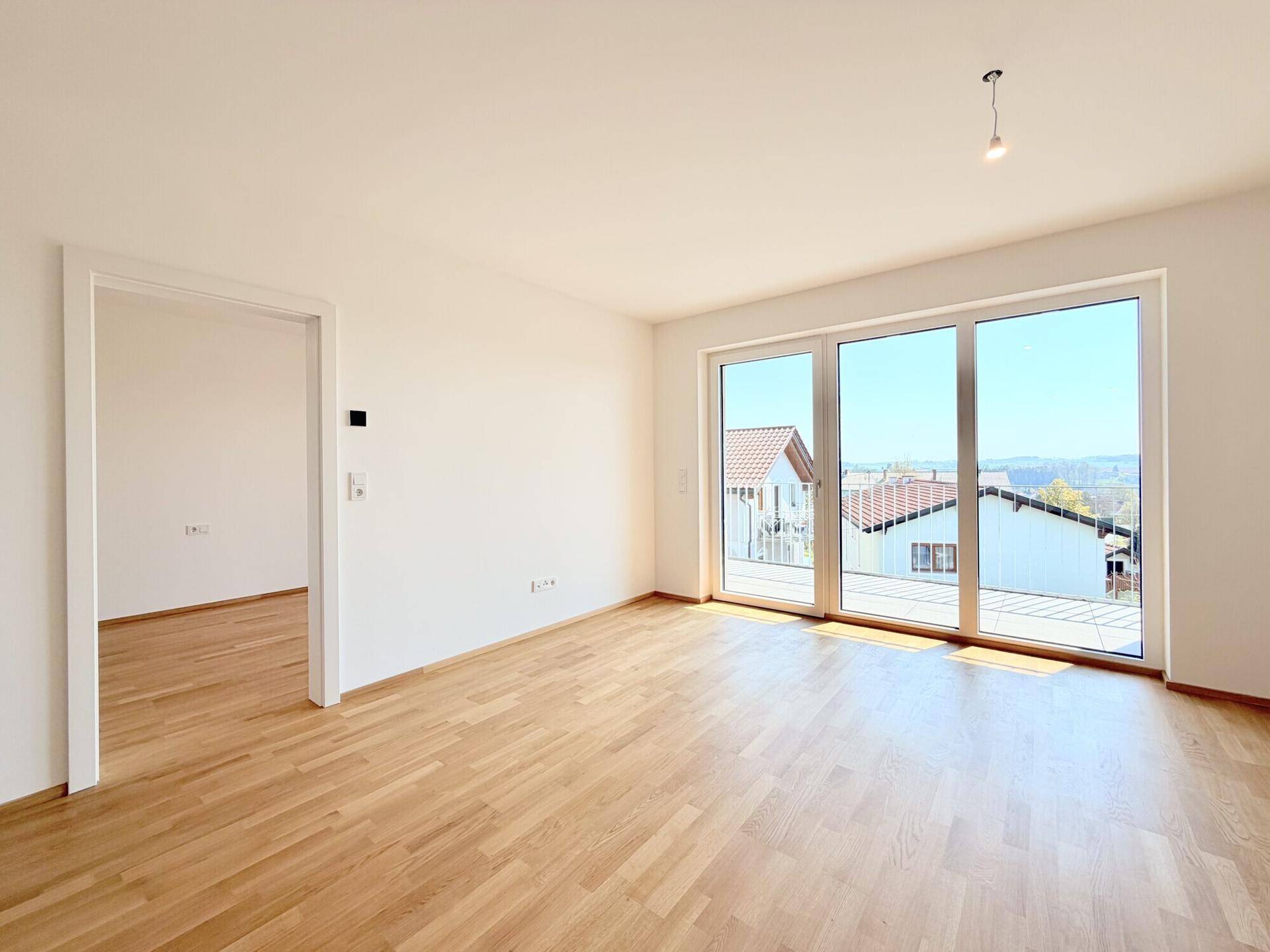 Heller Raum mit Holzboden und großem Fenster, das viel Licht spendet und einen Zugang zu einem weiteren Zimmer ermöglicht.