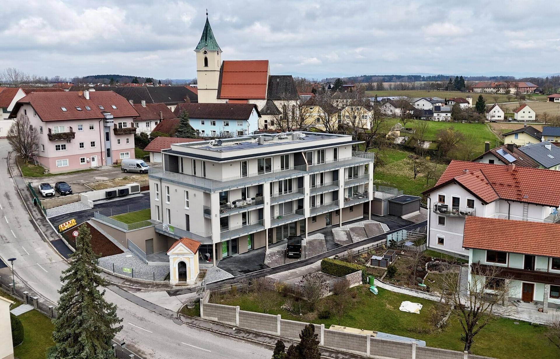 Luftaufnahme eines neu errichteten Mehrfamilienhauses in ländlicher Umgebung mit Kirchturm im Hintergrund.