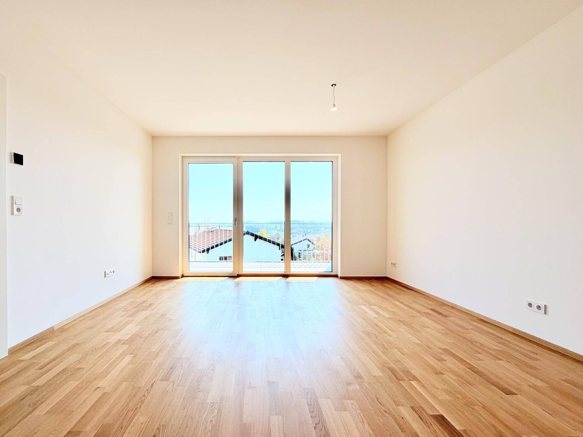 Helles, leeres Zimmer mit Holzboden und großen Fenstern, die viel Tageslicht hereinlassen.