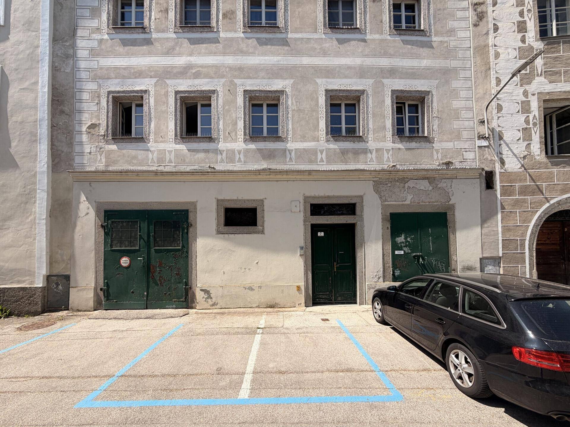 Historische Gebäudefassade mit mehreren Fenstern, zwei grünen Türen im Erdgeschoss und einem Parkplatz davor.