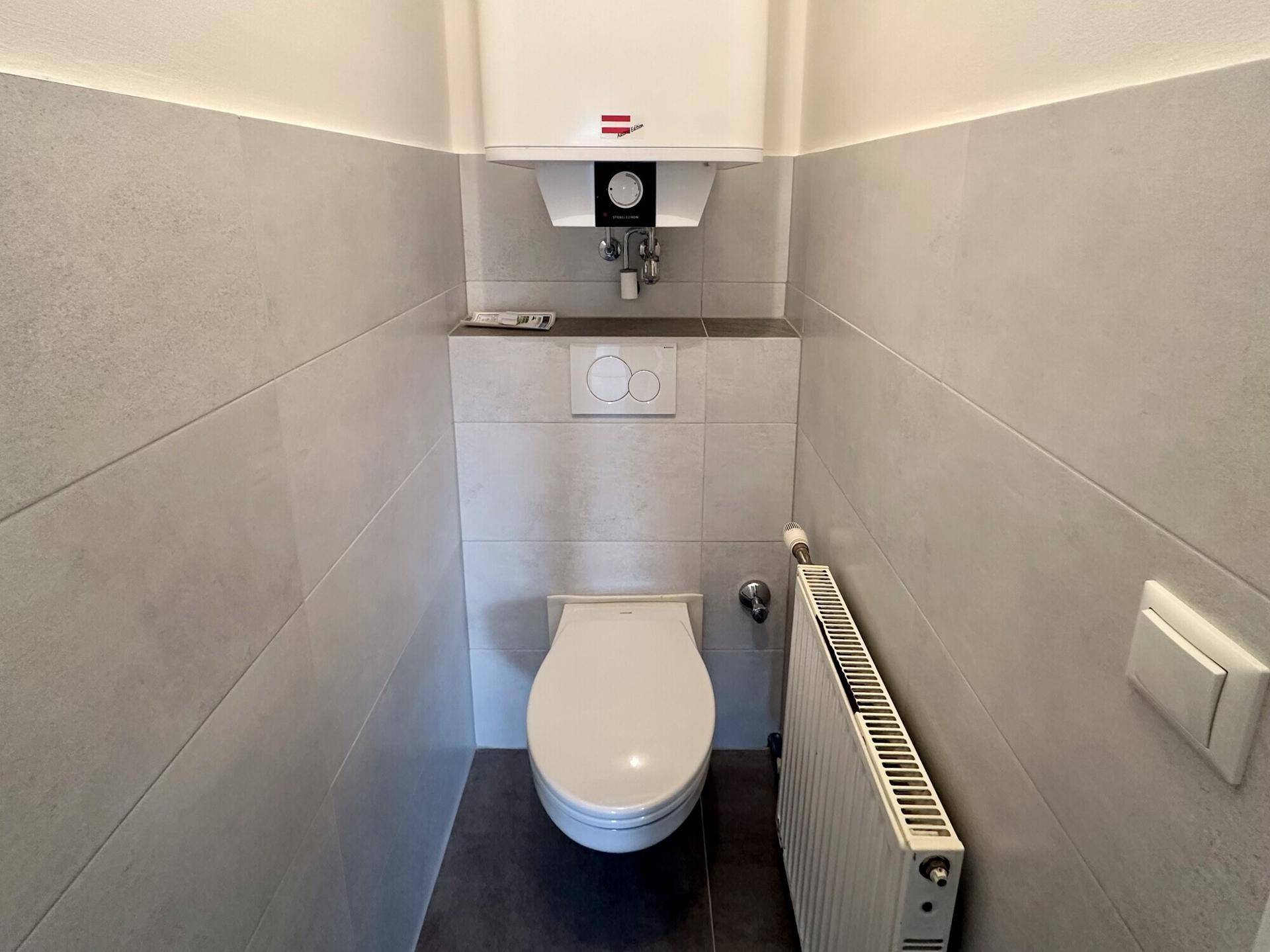 Separates WC mit wandhängender Toilette, Heizkörper und modern gefliesten Wänden.