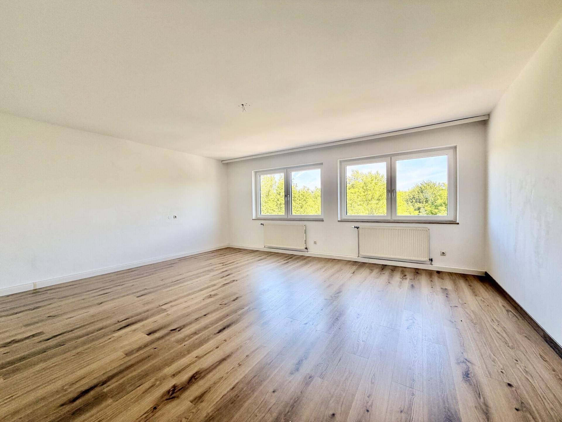 Lichtdurchflutetes Zimmer mit modernem Laminatboden und Ausblick auf grüne Bäume.