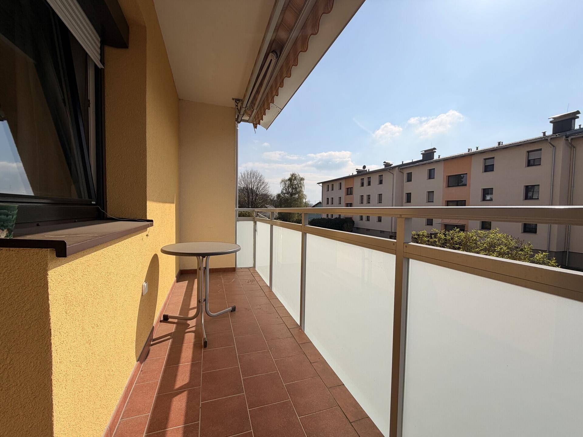 Sonniger Balkon mit Terrakottafliesen, kleinem Tisch und Blick auf die Nachbarschaft.
