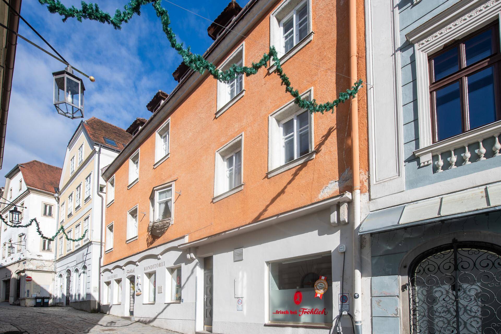 Ansicht des historischen Gebäudes mit oranger Fassade und Geschäften im Erdgeschoss an einer belebten Gasse.