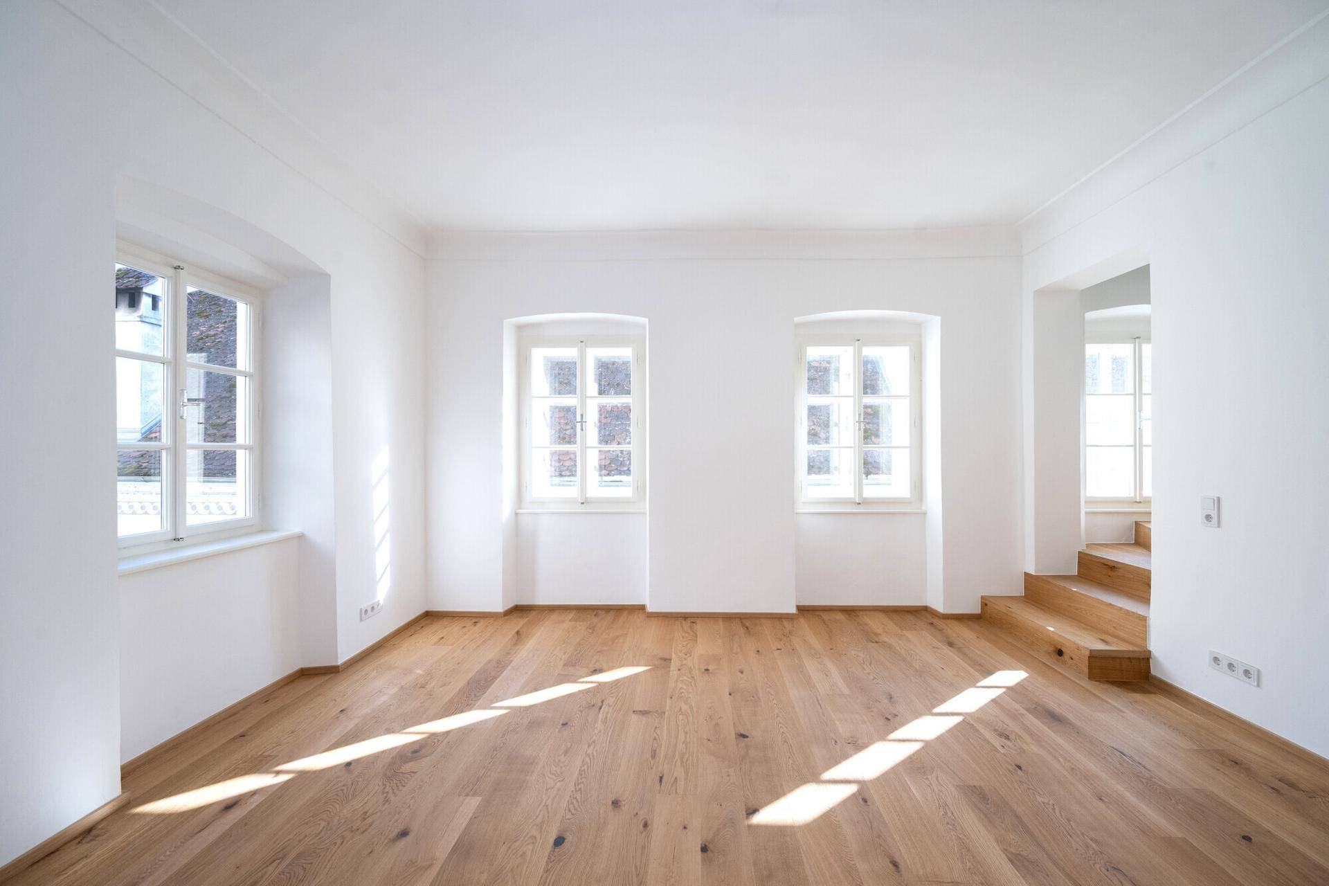 Lichtdurchfluteter Raum mit Holzboden und mehreren Fenstern, ideal als Wohnbereich.